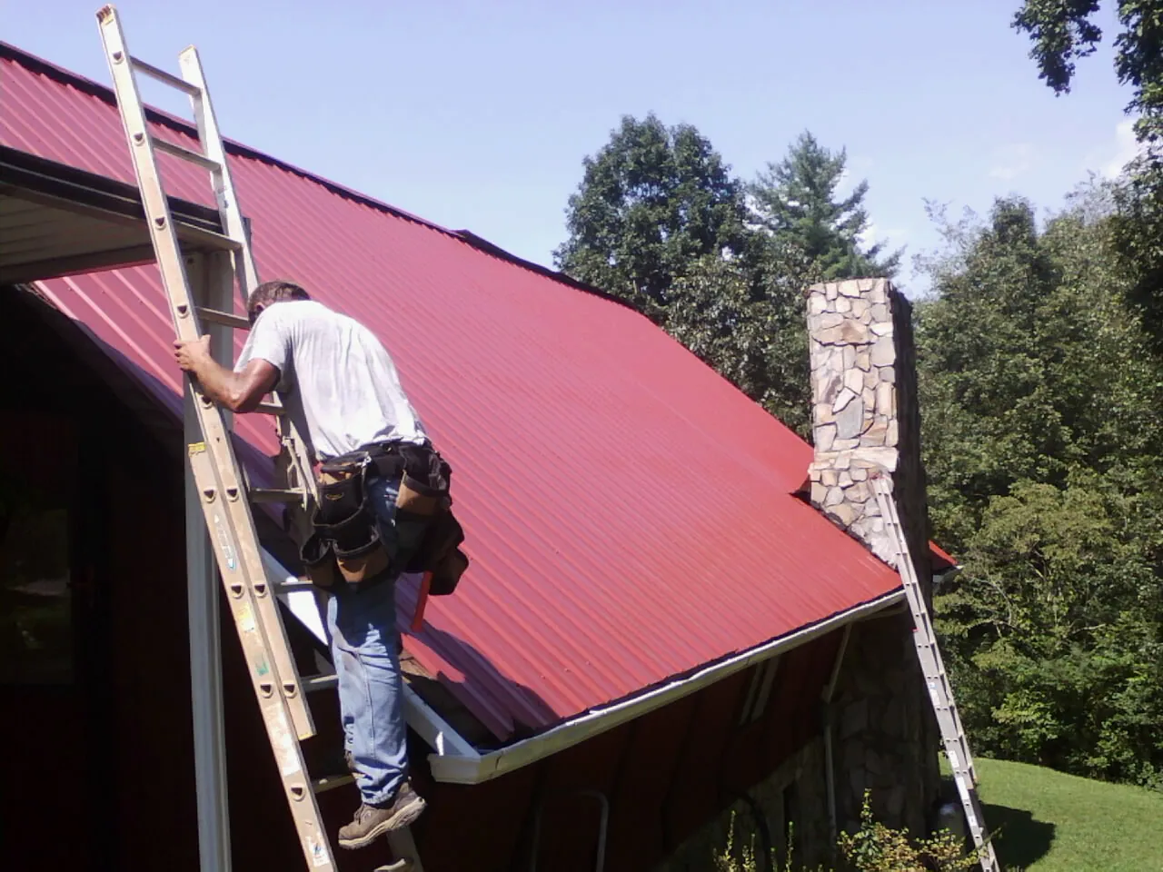 Roofer on a ladder beside a red metal roof and stone chimney — Soffit & Fascia Repair by Roofing Brothers in Barre